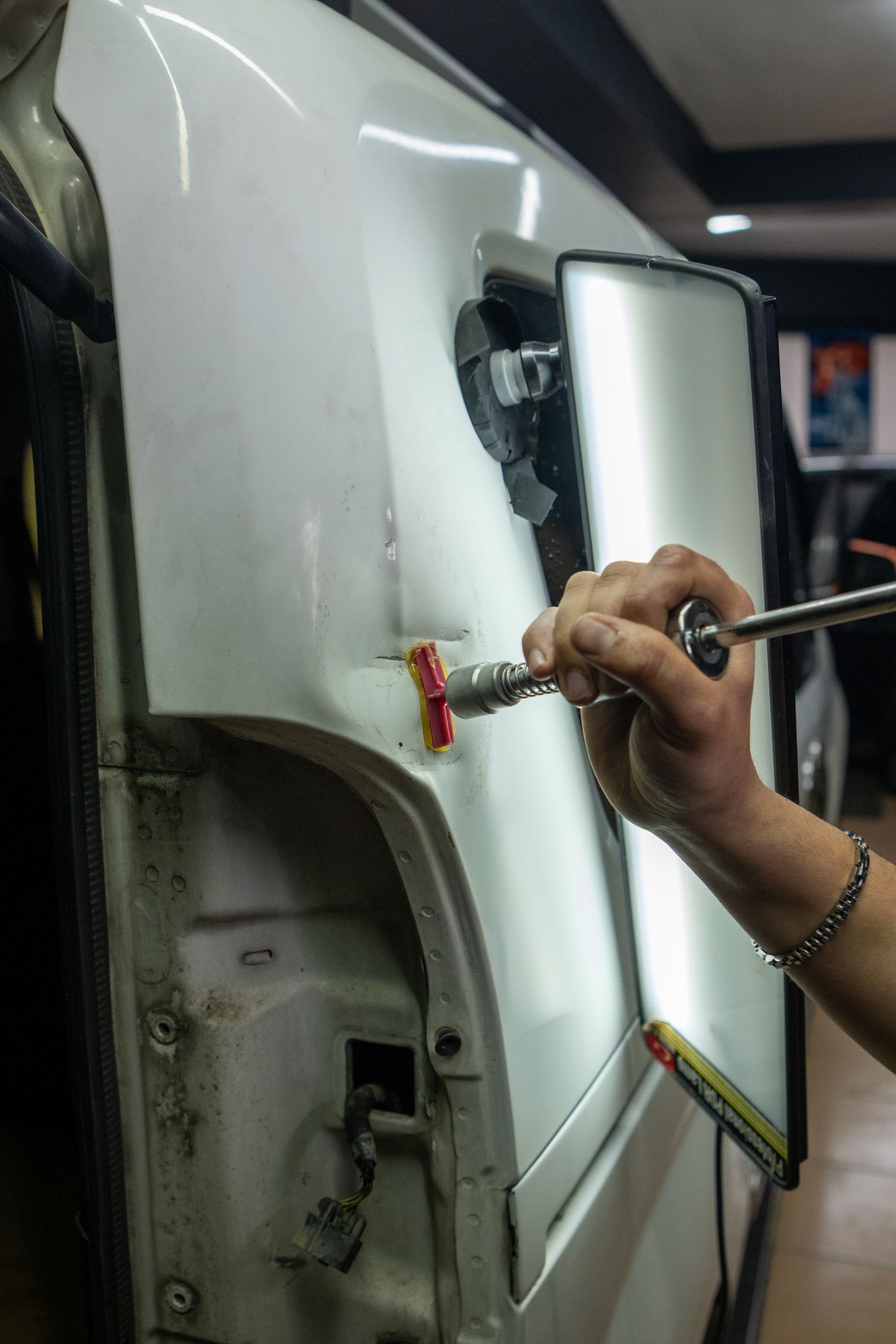 Technician lifting a dent from a white door panel using a glue-pull tab and a reflector board